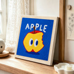 Bitten apple with big eyes punch needle artwork displayed on a table