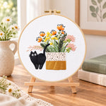 Black cat and wildflowers embroidery kit finished hoop displayed on a wooden stand as cozy tabletop decor