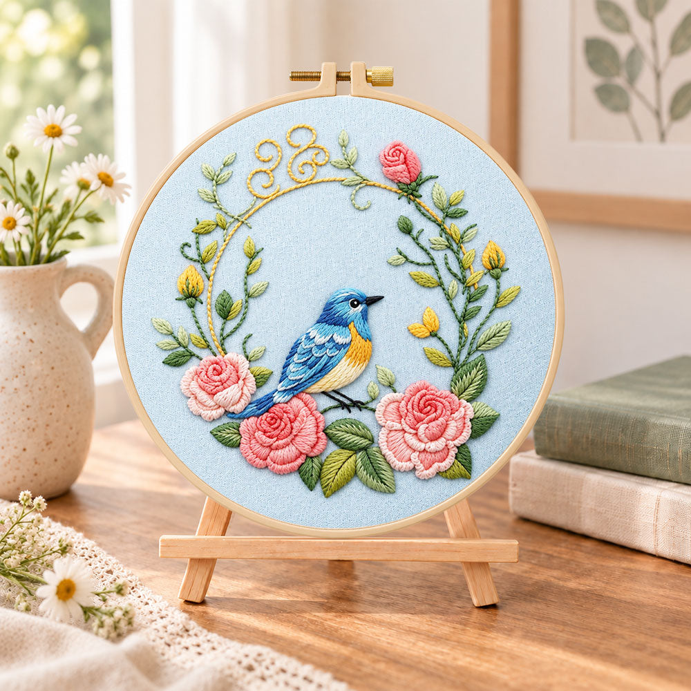 Blue bird inside pink rose wreath embroidery kit finished hoop displayed on a wooden stand as cozy tabletop decor