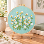 Daisies on teal fabric embroidery kit finished hoop displayed on a wooden stand as cozy tabletop decor