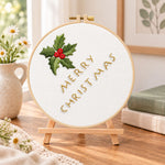 Holly and merry christmas embroidery kit finished hoop displayed on a wooden stand as cozy tabletop decor
