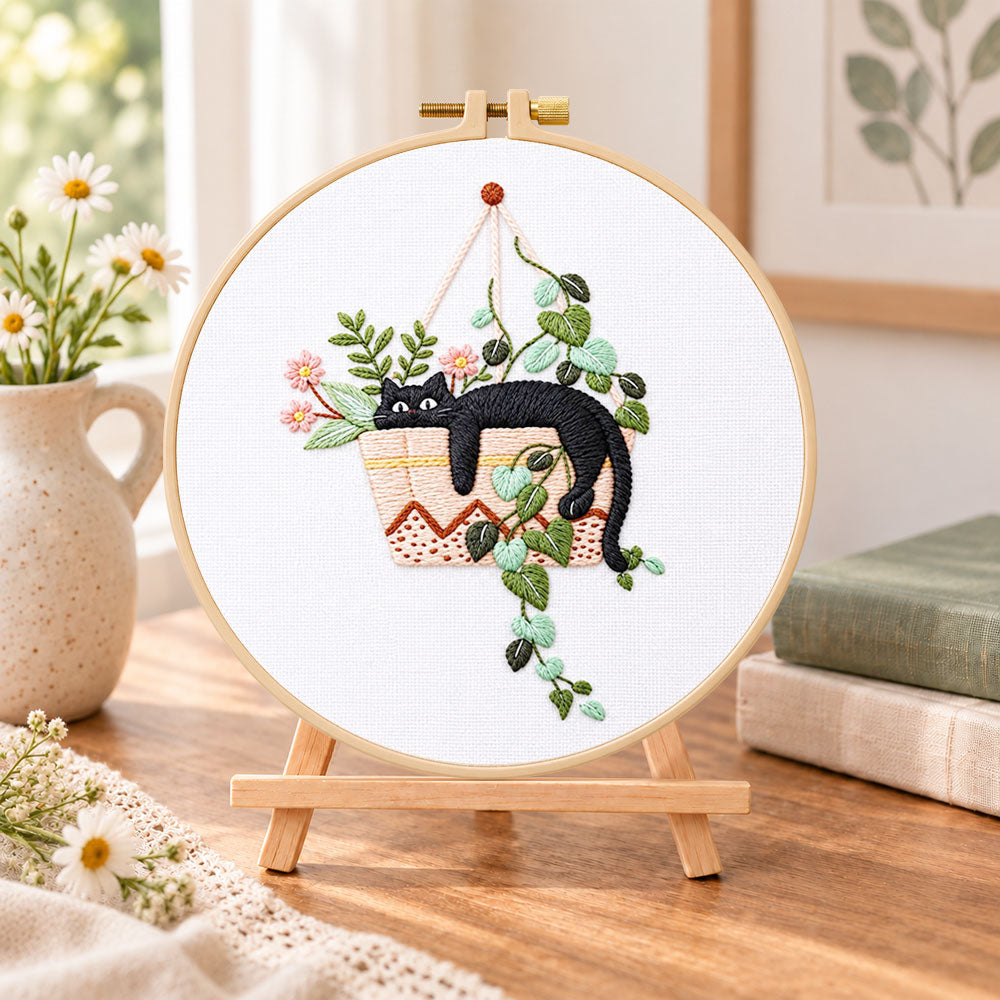 Lazy cat in hanging planter embroidery kit finished hoop displayed on a wooden stand as cozy tabletop decor
