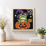 Finished little witch frog sitting on pumpkin artwork displayed on a wall