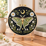 Luna moth and crescent moon embroidery kit finished hoop displayed on a wooden stand as cozy tabletop decor