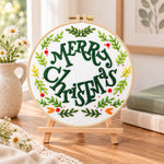 Merry christmas wreath embroidery kit finished hoop displayed on a wooden stand as cozy tabletop decor