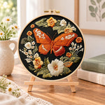 Orange butterfly amid blooms embroidery kit finished hoop displayed on a wooden stand as cozy tabletop decor