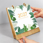 Hands flipping the botanical leaf cover page on a wooden desk calendar with gold metal binder rings