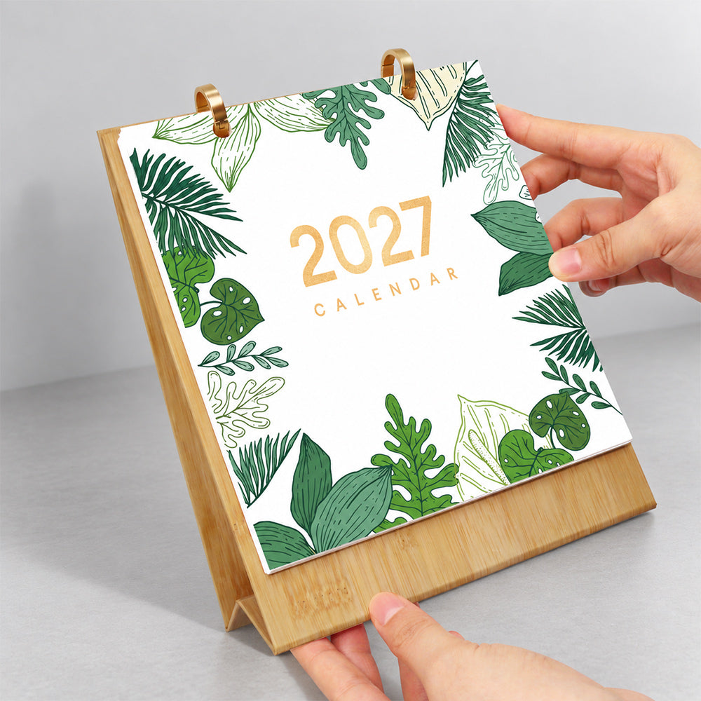 Hands flipping the botanical leaf cover page on a wooden desk calendar with gold metal binder rings