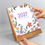 Hands flipping the floral cover page on a wooden desk calendar with gold metal binder rings