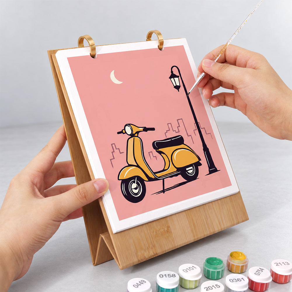 Hands painting a yellow vintage scooter illustration on the On the Move Paint by Numbers Desk Calendar
