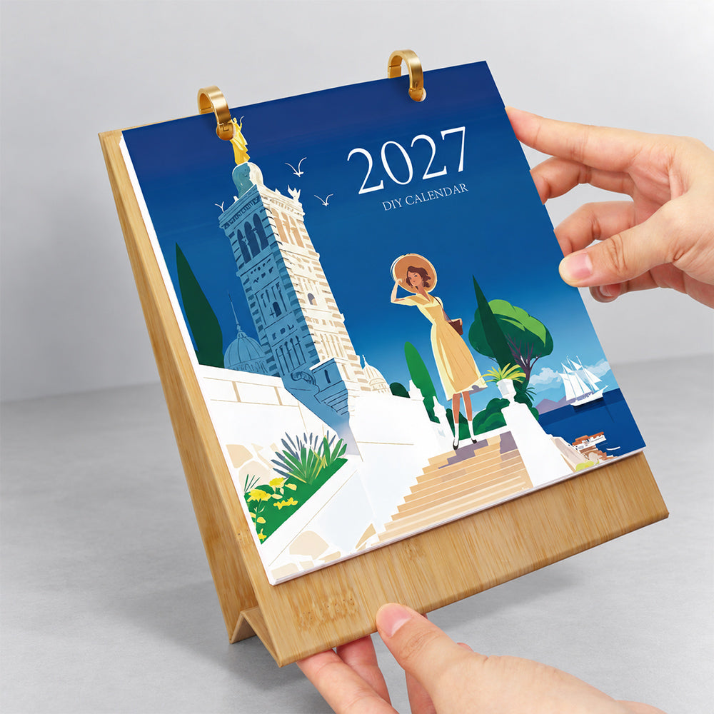 Hands flipping the travel-themed cover page on a wooden desk calendar with gold metal binder rings