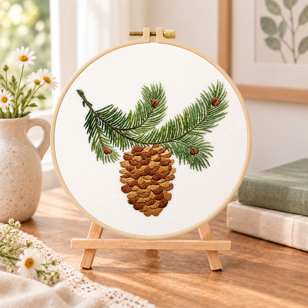 Pine branch and cone embroidery kit finished hoop displayed on a wooden stand as cozy tabletop decor