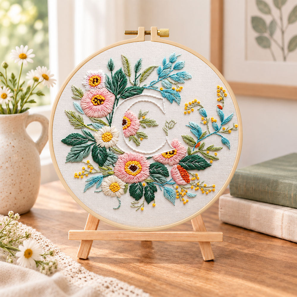 Pink botanical blooms and letter c embroidery kit finished hoop displayed on a wooden stand as cozy tabletop decor