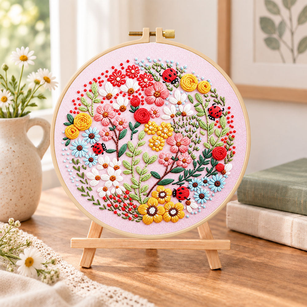 Pink floral art with ladybugs embroidery kit finished hoop displayed on a wooden stand as cozy tabletop decor
