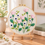 Pink thistle and daisies embroidery kit finished hoop displayed on a wooden stand as cozy tabletop decor