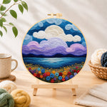 Finished purple clouds and flower-covered coastline needle felting displayed on a wooden easel