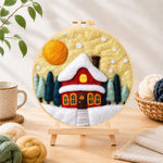 Finished red cottage under snowy yellow sky needle felting displayed on a wooden easel