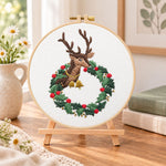 Reindeer in holly wreath embroidery kit finished hoop displayed on a wooden stand as cozy tabletop decor