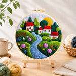 Finished serene blue river and red roof village needle felting displayed on a wooden easel