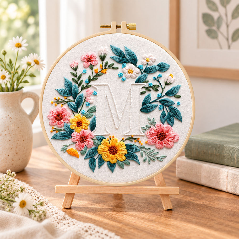 Sunny floral wreath and letter m embroidery kit finished hoop displayed on a wooden stand as cozy tabletop decor