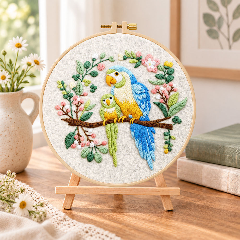 Two parrots on blossom branch embroidery kit finished hoop displayed on a wooden stand as cozy tabletop decor