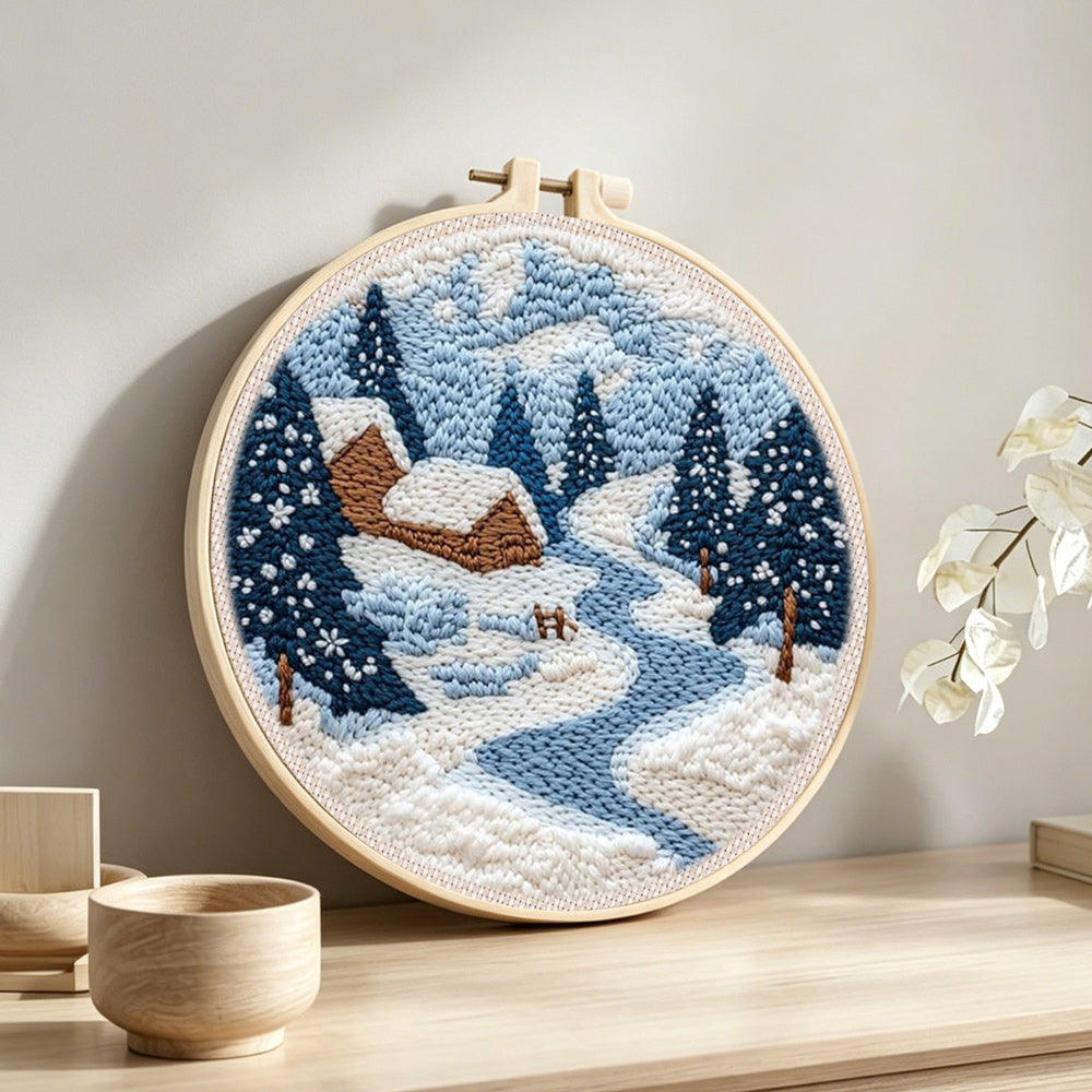 Winter cabin scene with snowy pine trees and blue river punch needle artwork displayed on a table
